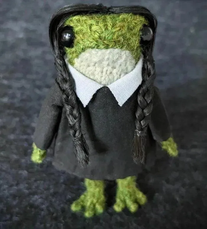 a handmade crocheted frog with green textured skin, wearing a black dress with a white collar, and has long black braids…