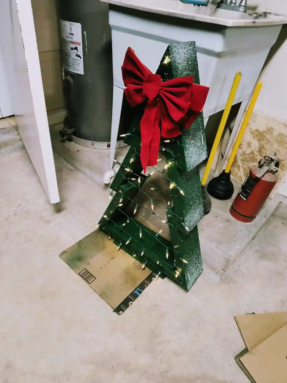 A small, glittery green cardboard Christmas tree with string lights and a large red bow stands on a cardboard box in a…