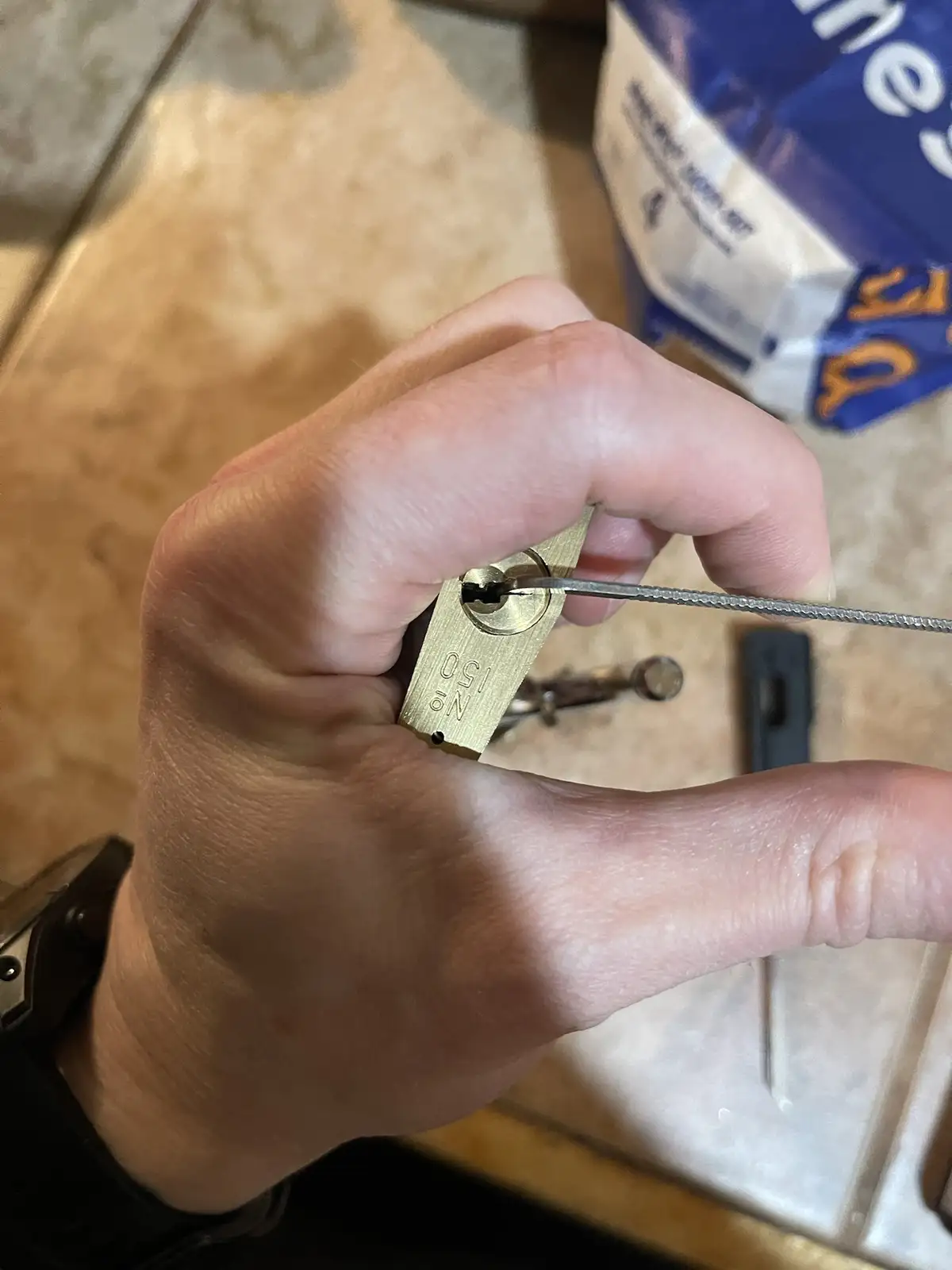 A close-up photo shows a person's hand holding a brass padlock while inserting a thin, serrated metal tool into its keyhole…
