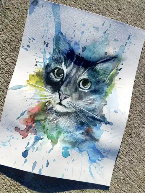 A watercolor painting of a cat's face on white paper, surrounded by colorful paint splatters in blue, green, and red, lying…