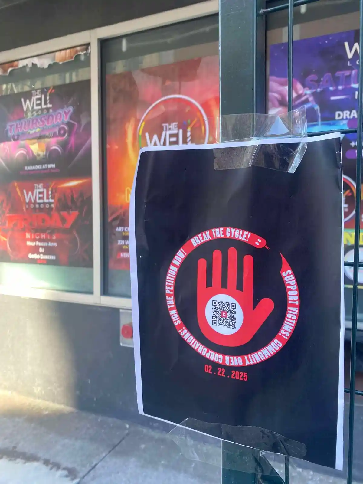 A black poster taped to a metal gate in front of a club called 'The Well London' features a large red handprint with a QR…