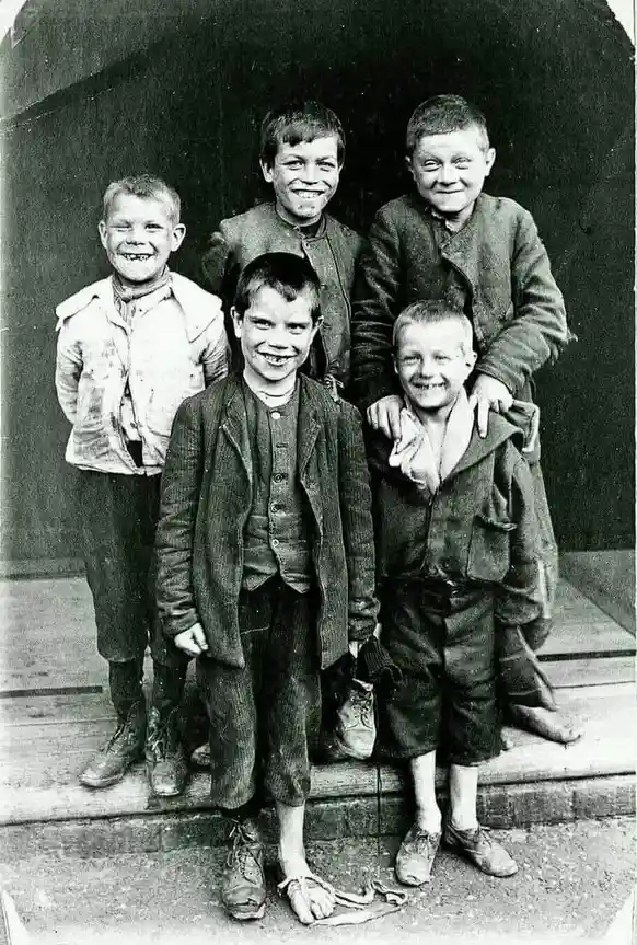 A vintage black-and-white photograph shows five young boys posing on wooden steps, all smiling broadly despite their worn…