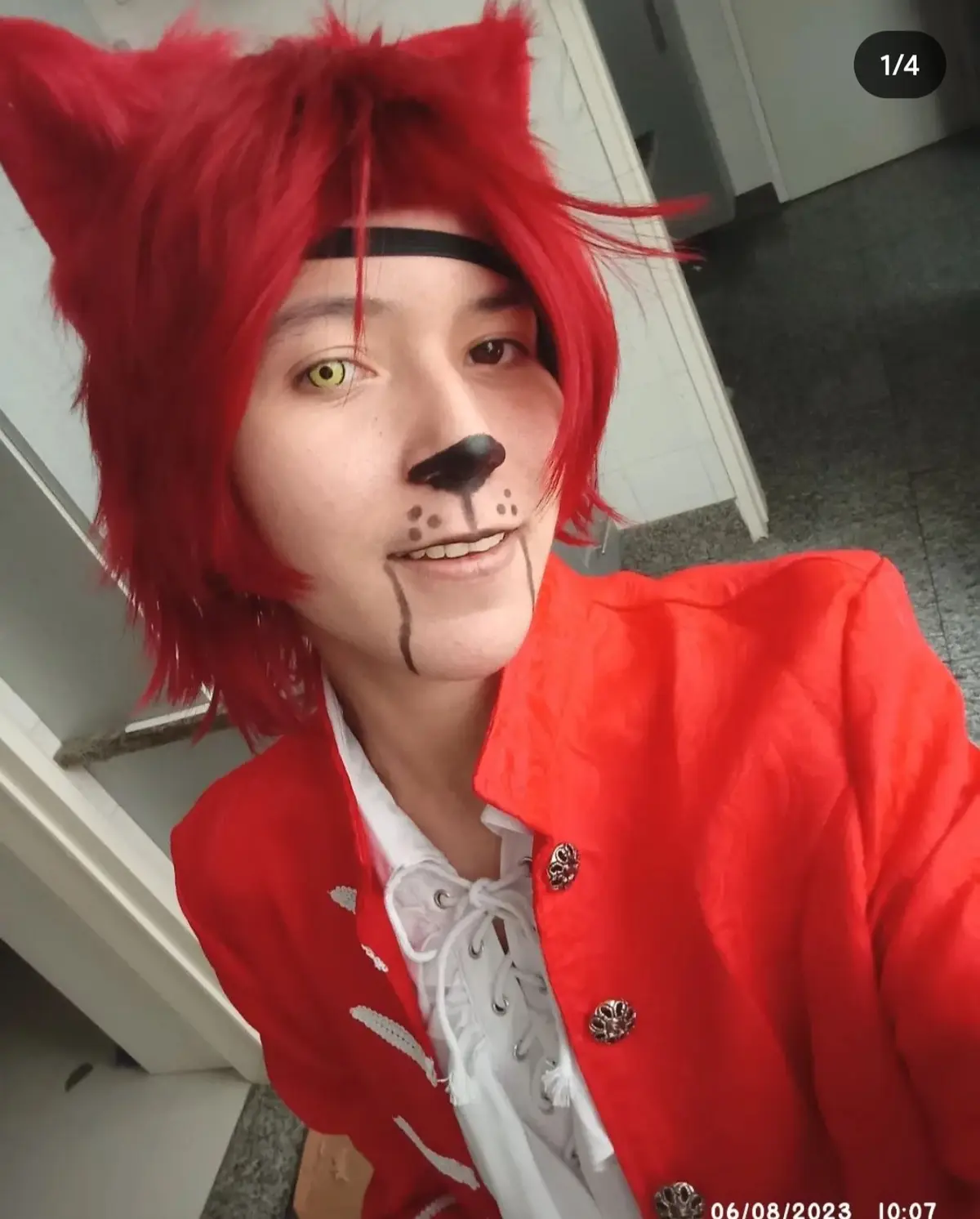 A person in a selfie poses with bright red wig, red fox ears, and fox face paint including a nose and whisker markings…