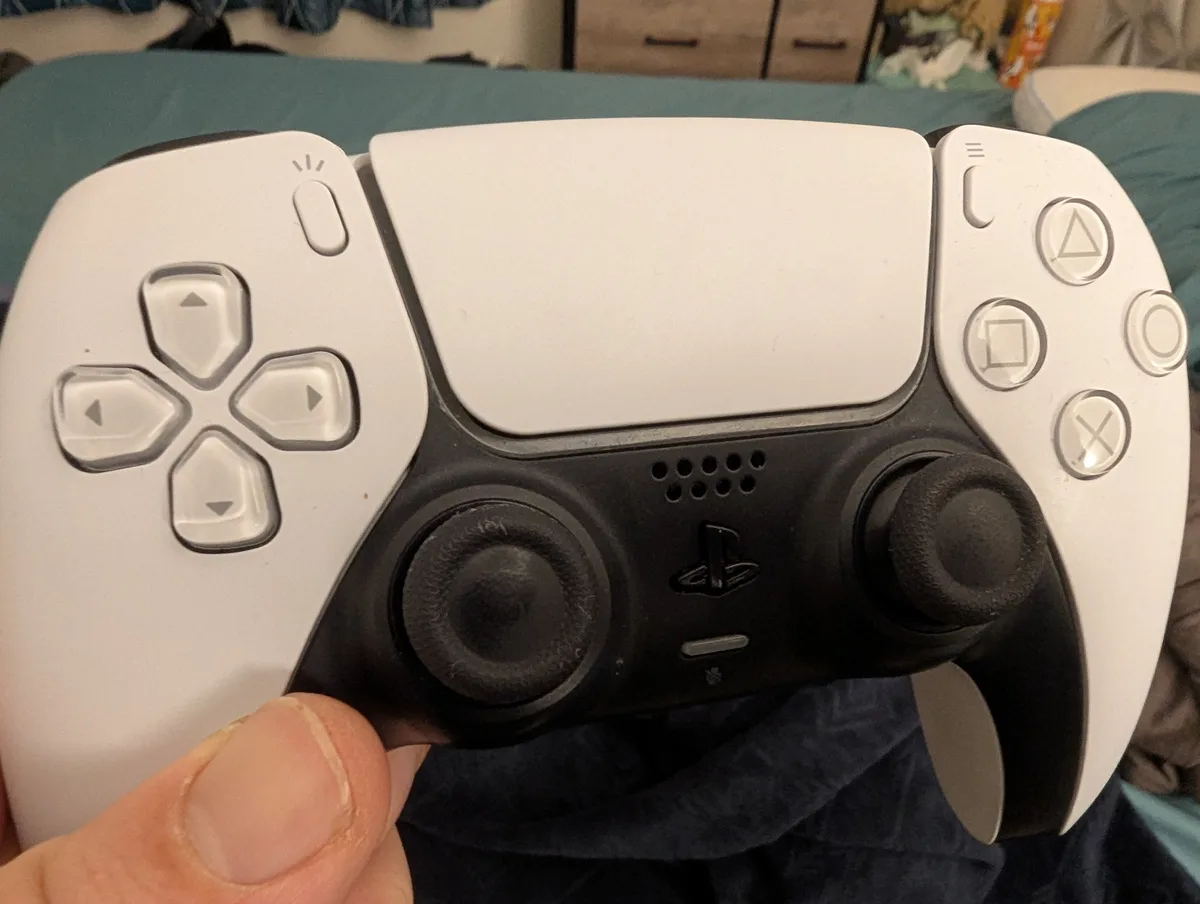 A close-up photo of a white and black PlayStation 5 DualSense controller being held in a person's hand, showing the…