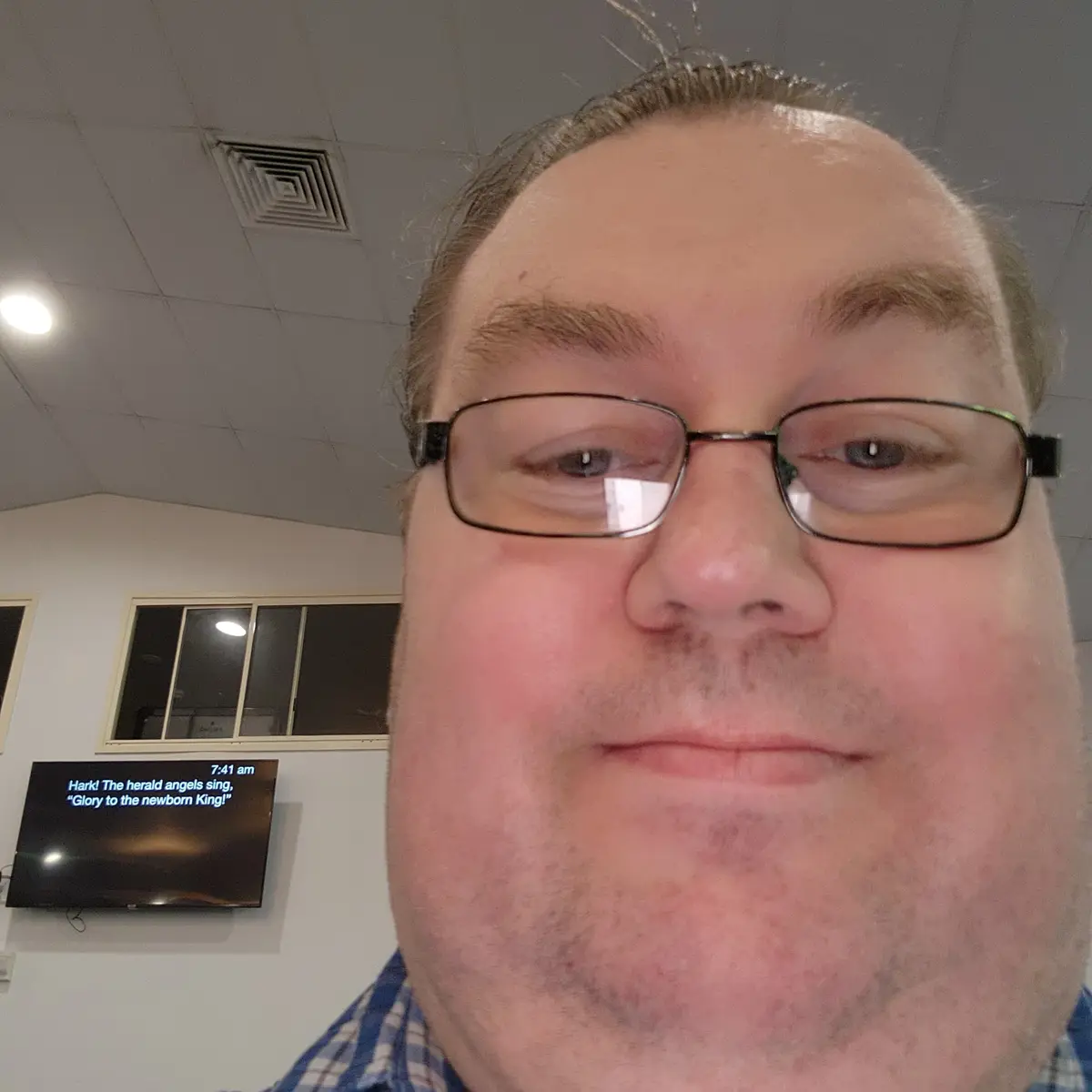A close-up selfie of a man with glasses and a slight smirk, taken indoors in front of a wall-mounted TV displaying the…