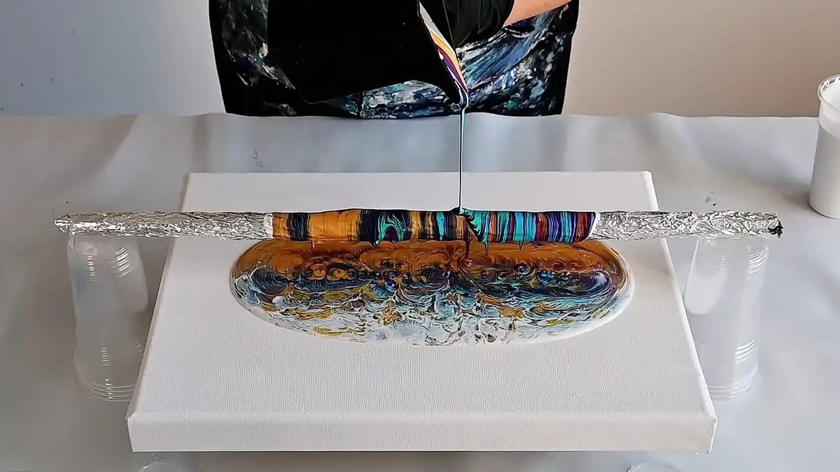 A person pours colorful, viscous paint onto a white canvas that is supported by aluminum foil-covered sticks resting on…