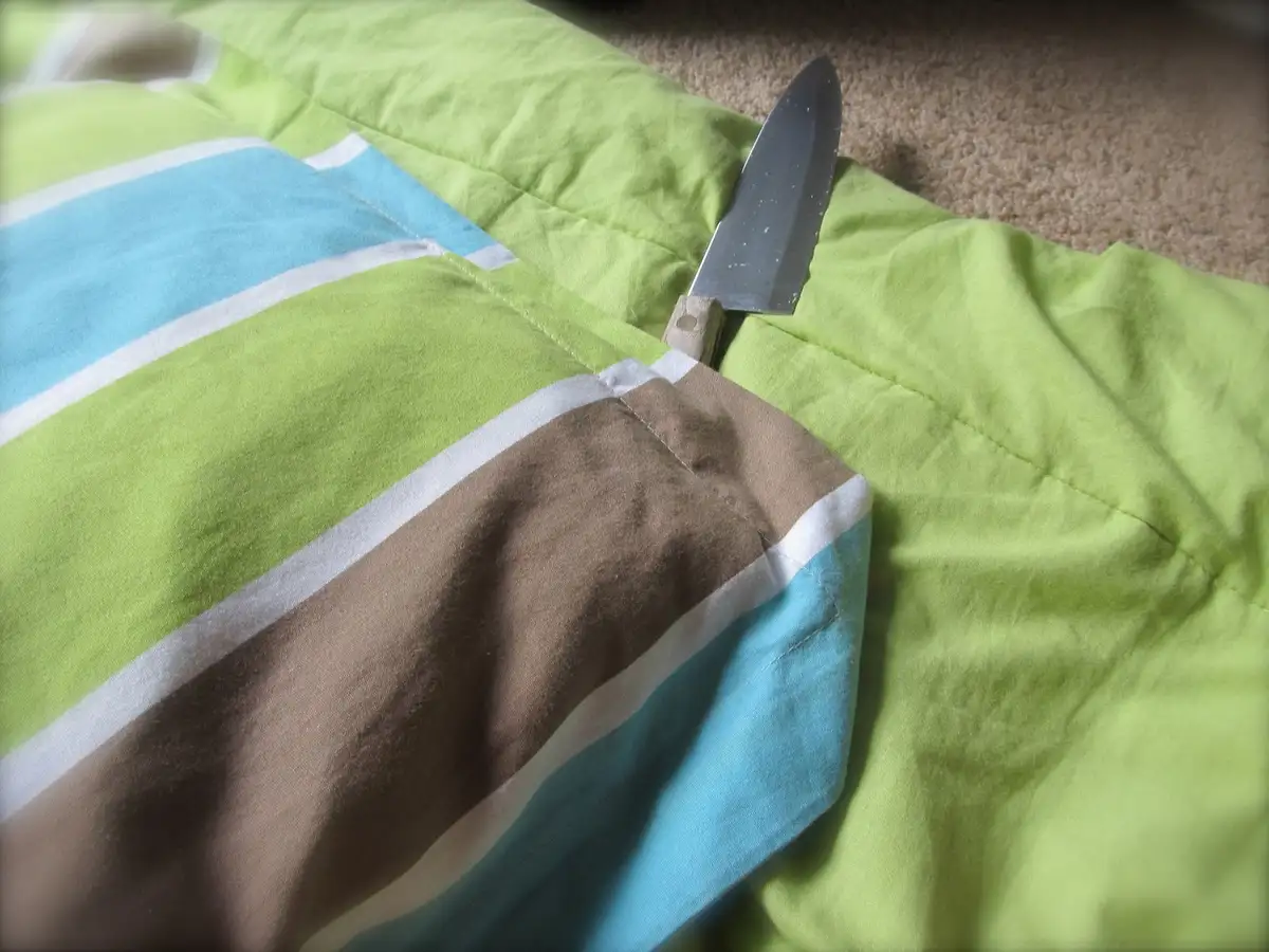A close-up photo shows a small knife with a wooden handle stuck into the seam of a brightly colored striped pillow, which…