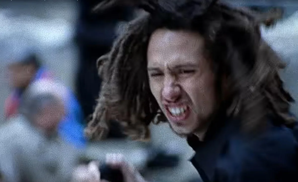 A man with long dreadlocks is shown in extreme close-up, screaming with his eyes squeezed shut and teeth bared, conveying…