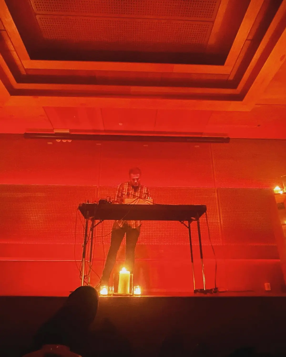 A person stands behind a keyboard on a stage bathed in intense red lighting, with several lit candles placed at the front of…
