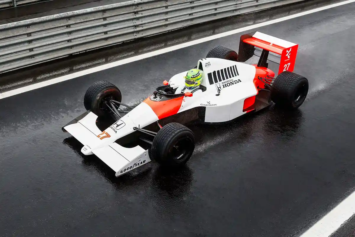 A white and orange Formula 1 race car with the number 27, sponsored by Honda and Goodyear, drives on a wet racetrack. The…