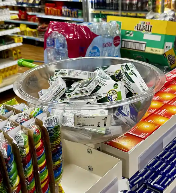 A clear plastic bowl in a supermarket aisle is filled with small boxes of 'Allergy Relief' tablets, priced at £1 each. The…
