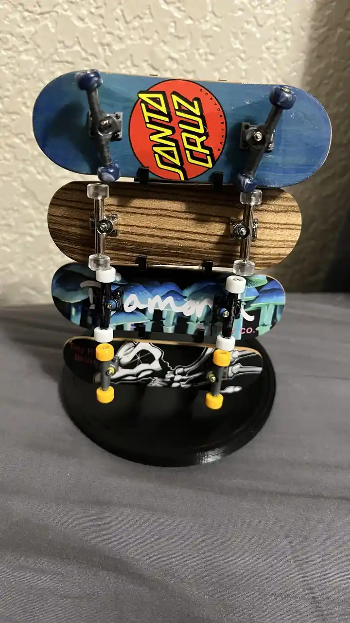 A vertical stack of four miniature finger skateboards displayed on a black stand, each with different graphics: top has a…