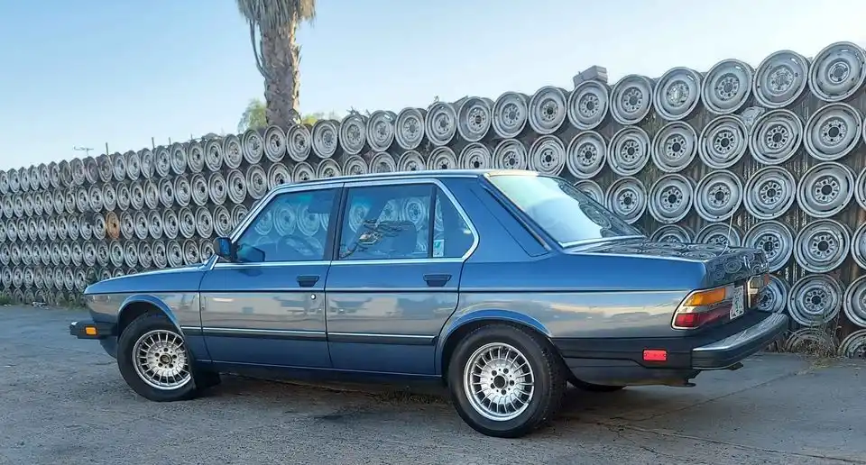 A blue BMW sedan is parked in front of a wall made entirely of stacked wheels. The car has wheels on its own tires, and the…
