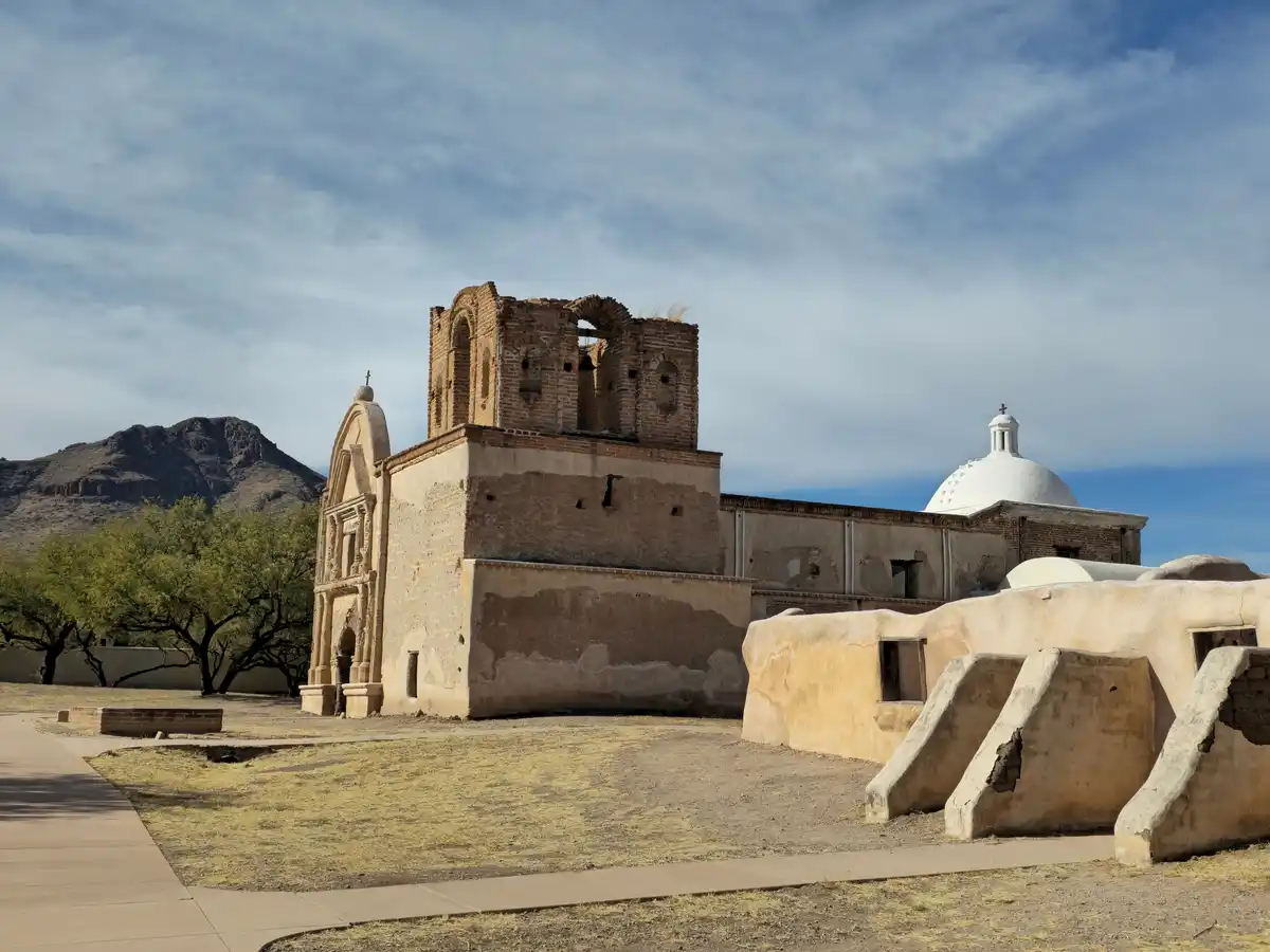 A photograph of a historic, partially ruined adobe church complex in a desert landscape with mountains in the background…