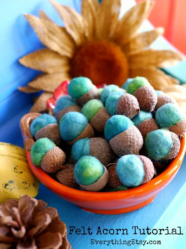 A bright orange bowl filled with handmade felt acorns, each with a brown textured cap and a blue-green felt body, sits on a…