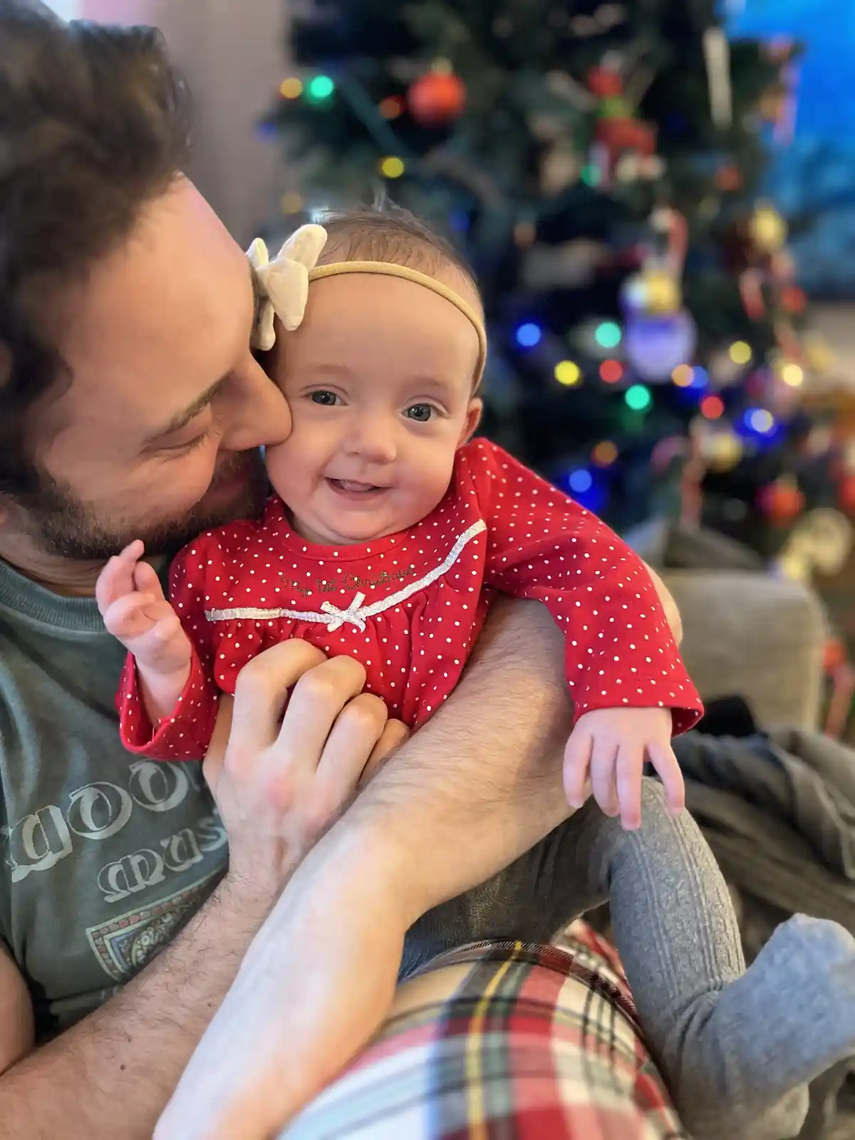 A man with a beard is holding a smiling baby girl in a red polka-dot outfit with a white bow headband. The baby's outfit has…