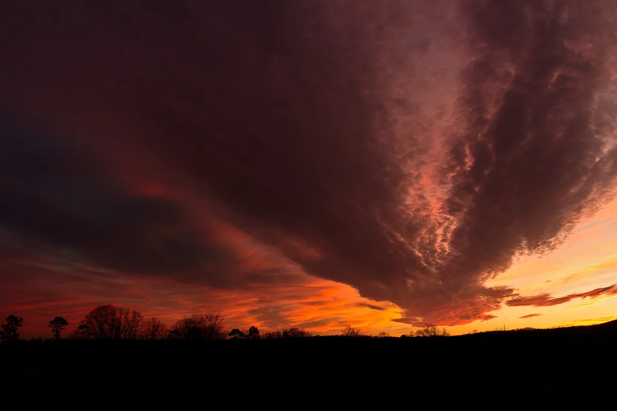 A dramatic sunset or sunrise photo showing a fiery orange and red sky with dark, textured clouds looming over a silhouette…