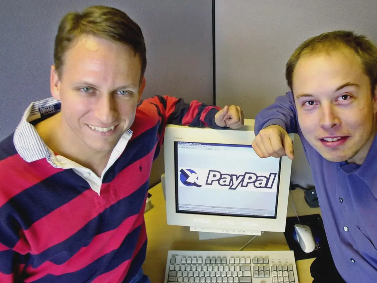 Two men, identified as PayPal co-founders, pose cheerfully beside an old beige computer monitor displaying the PayPal logo…