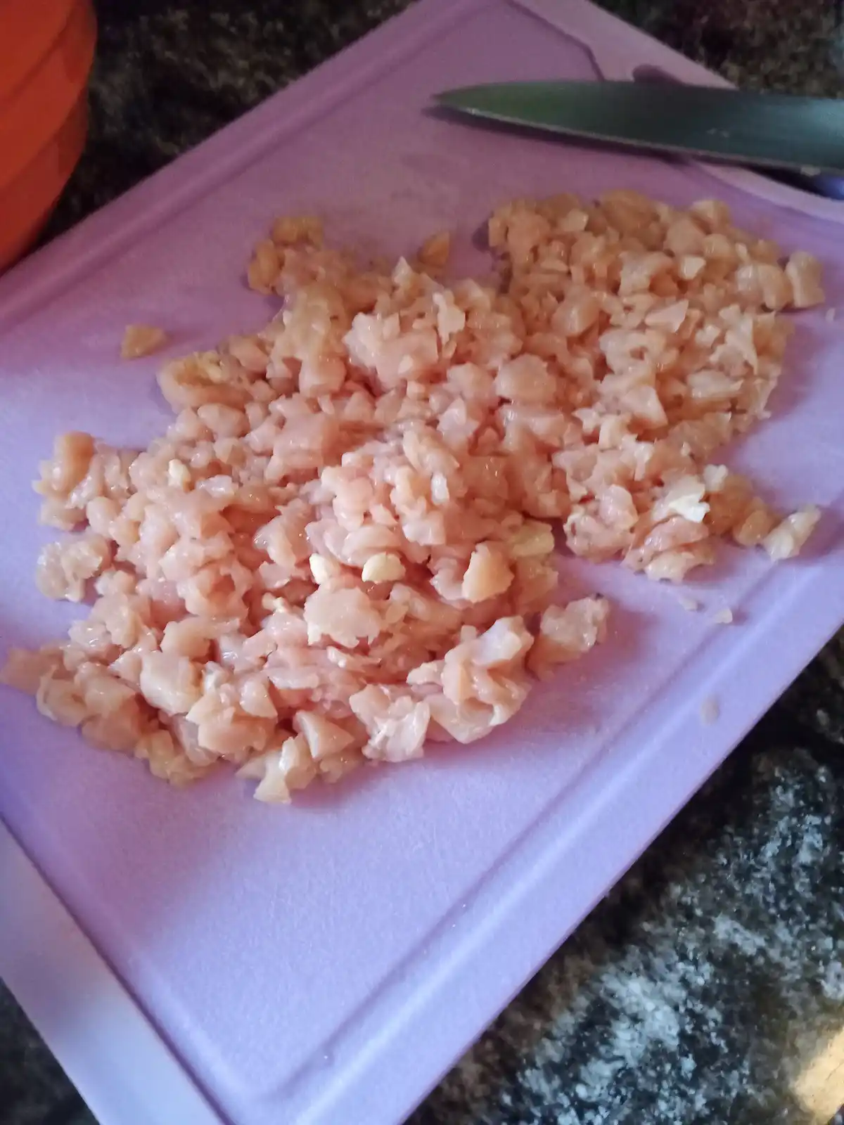 A close-up photo shows finely diced raw chicken meat arranged on a light purple plastic cutting board, with a knife resting…