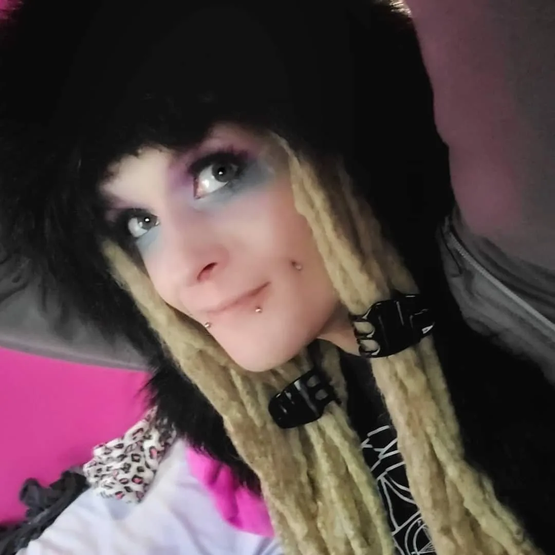A person with dramatic blue and purple eye makeup, blonde dreadlocks clipped with black plastic clips, and multiple facial…