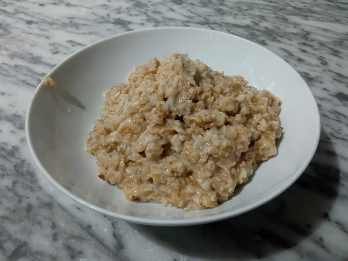A white bowl filled with plain, unadorned oatmeal sits on a gray and white marble countertop. The oatmeal appears simple and…