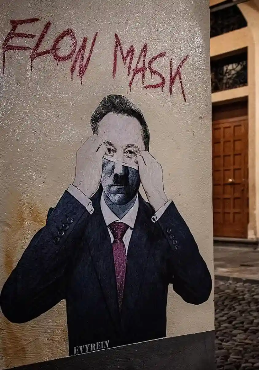 A street mural depicts a man in a suit and tie, with his hands pulling a white mask over his eyes, while red graffiti text…