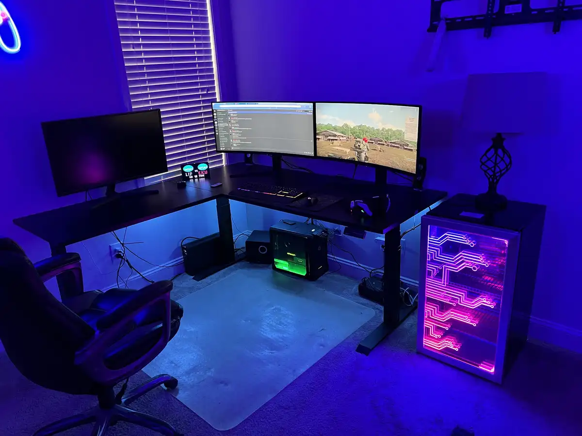 A dimly lit gaming room bathed in blue and purple LED lighting, featuring a large L-shaped desk with three monitors, one…