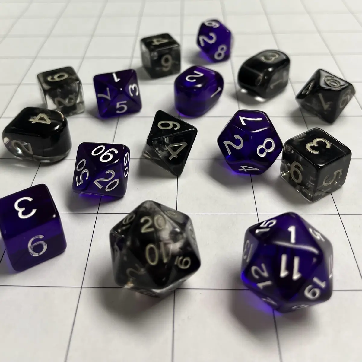 A collection of translucent purple and black polyhedral dice with silver numbers are scattered on a white gridded surface…