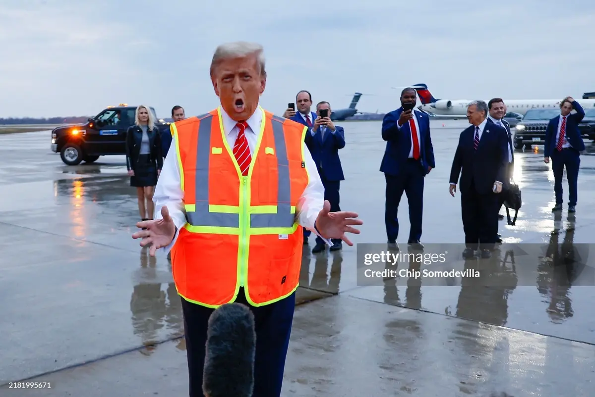 A man in a bright orange construction vest with reflective stripes stands on a wet airport tarmac, mouth open in an…