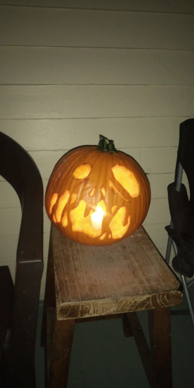 A carved jack-o'-lantern pumpkin with a lit candle inside sits on a wooden chair, its carved face glowing warmly. The…