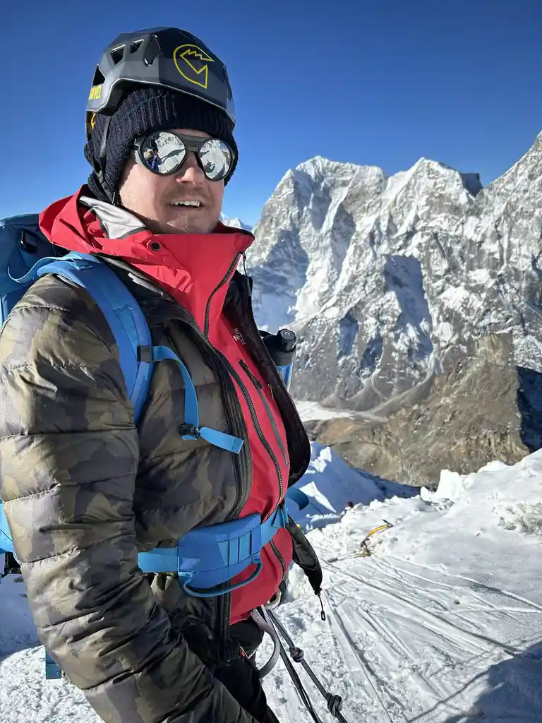 A man in full mountaineering gear, including a helmet, sunglasses, and a red and camouflage jacket, stands on a snowy…
