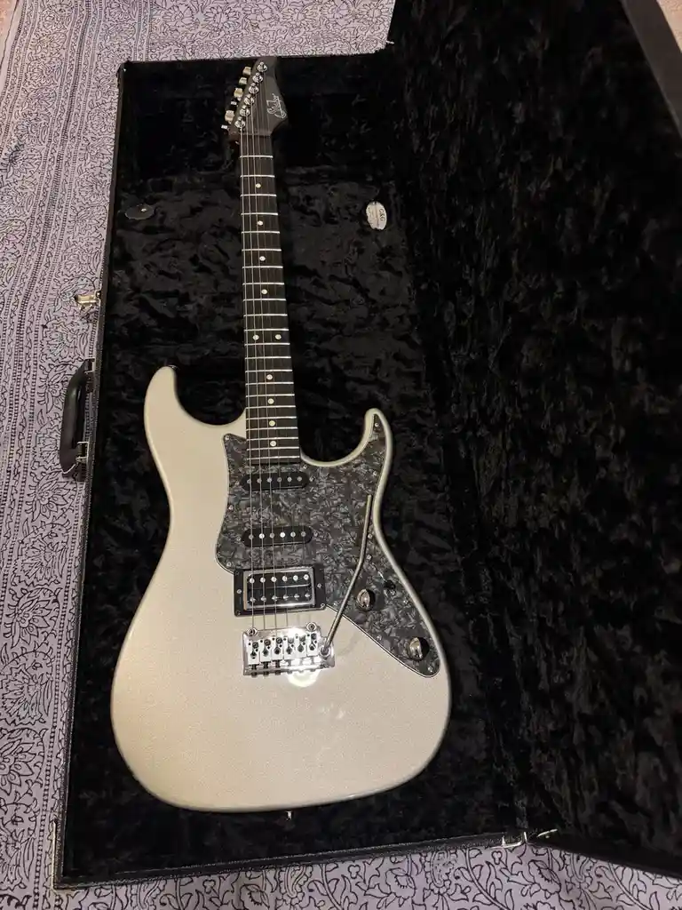 A silver sparkle electric guitar with a black pickguard and fretboard sits inside an open black velvet case. The case rests…
