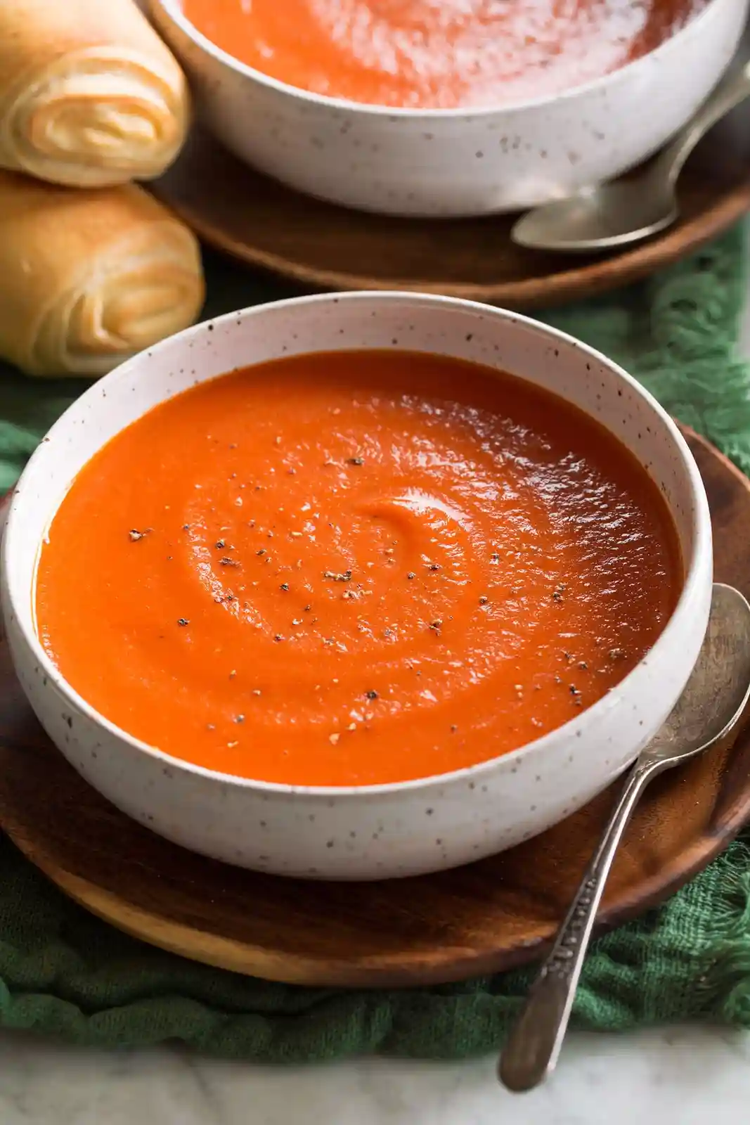 Two white speckled bowls filled with vibrant orange tomato soup, each with a spoon resting on a wooden plate, accompanied by…