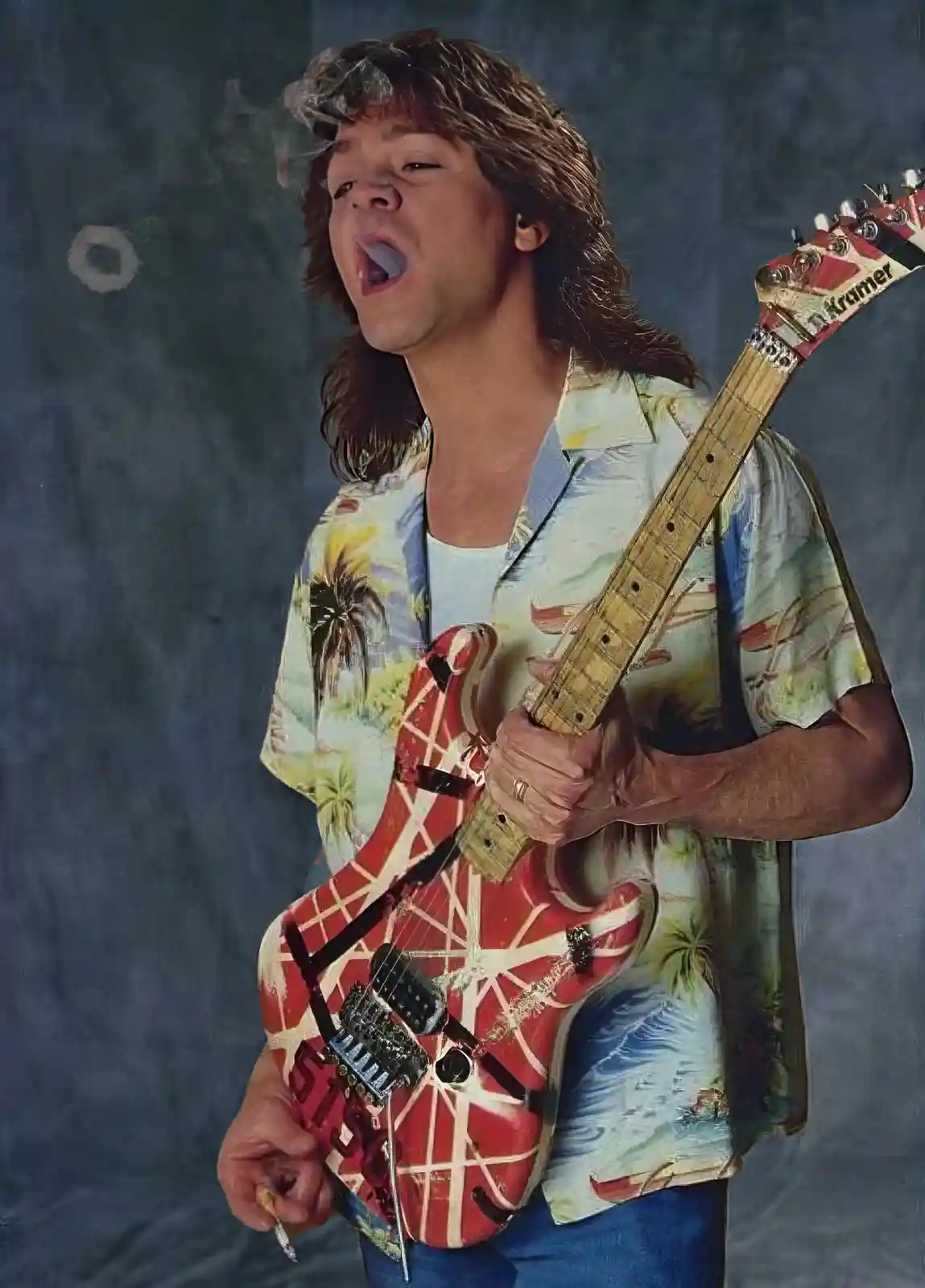 A man with long brown hair, wearing a tropical-print shirt and blue jeans, holds a red electric guitar with a white web…