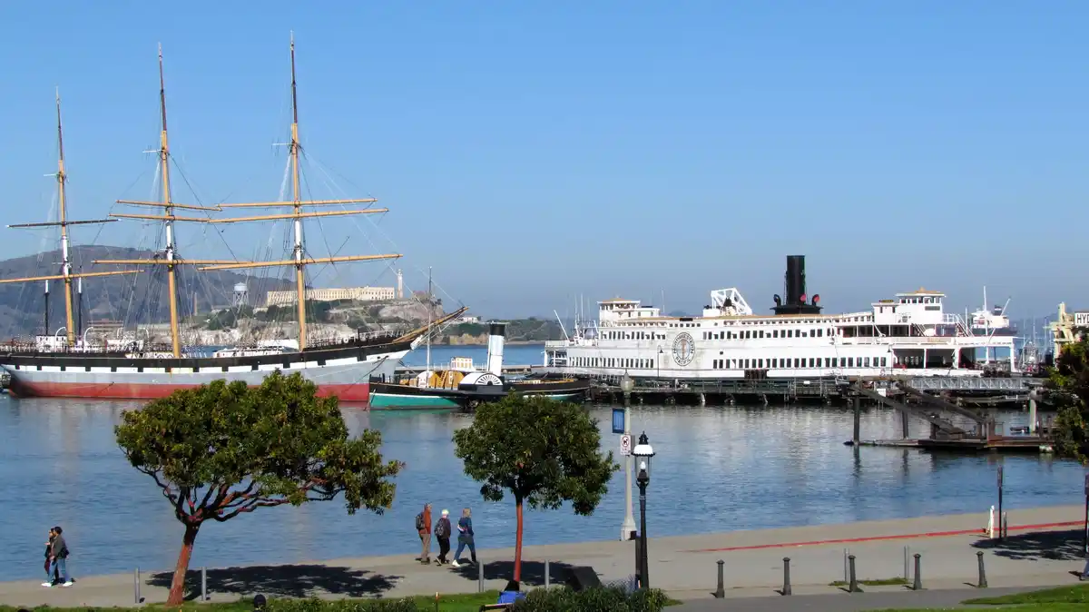A scenic harbor view shows a large, multi-masted sailing ship and a white steamship docked near Alcatraz Island. People walk…