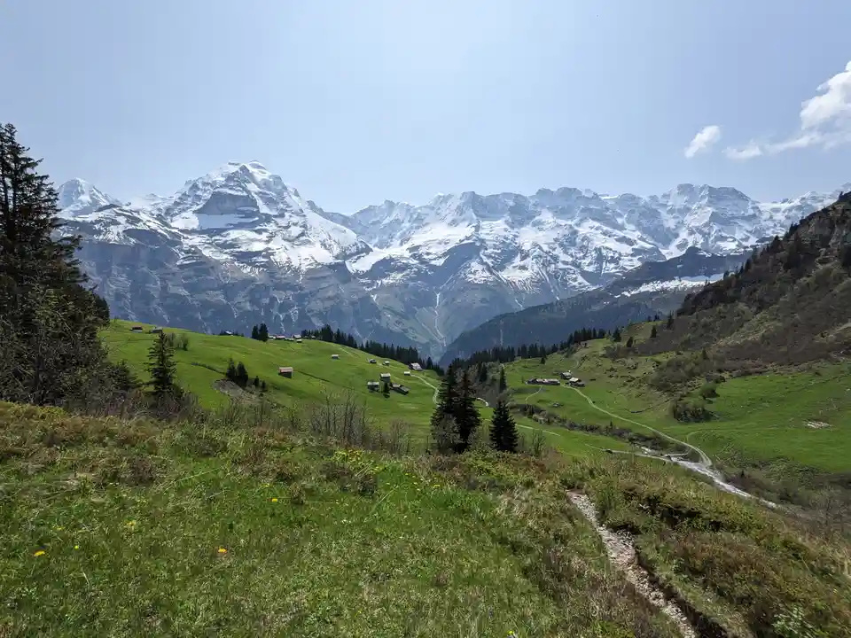 A scenic mountain landscape with snow-capped peaks in the background, green rolling hills in the midground dotted with small…