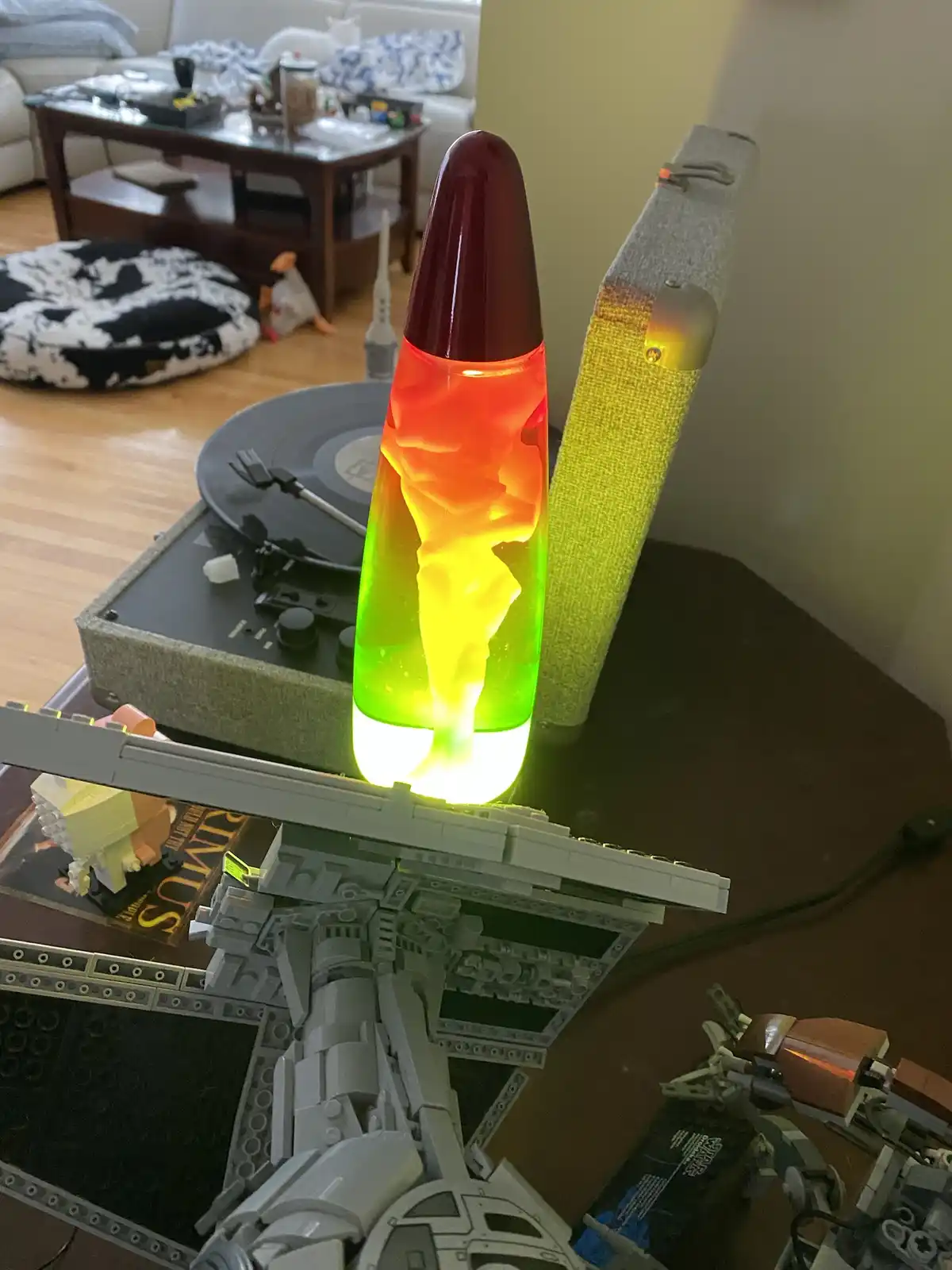 A close-up photo shows a colorful lava lamp with red, orange, and green wax, sitting on a table next to a gray LEGO Star…