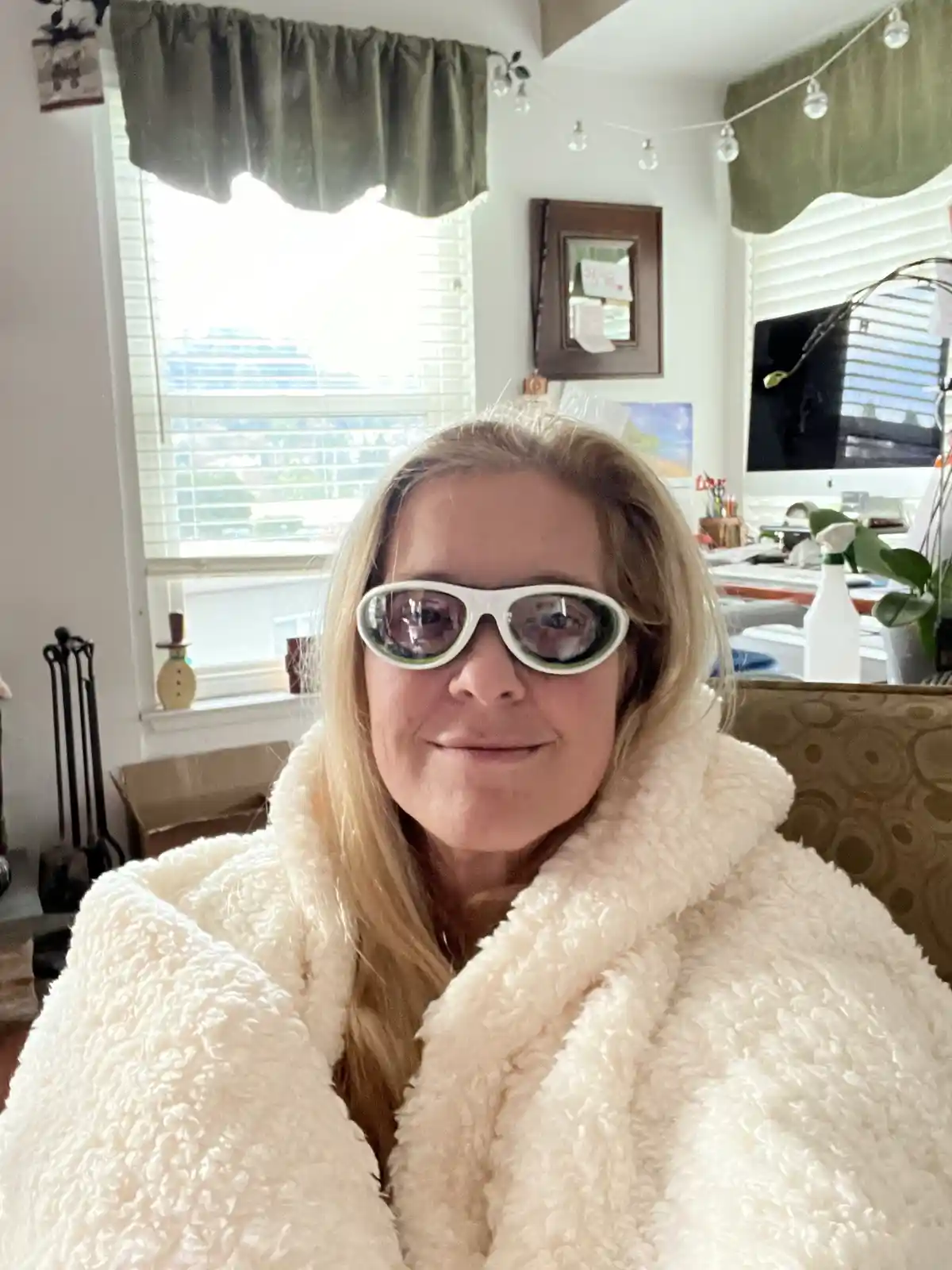 A woman with blonde hair wearing a fluffy white robe and oversized white-framed sunglasses sits in a cozy room with windows…