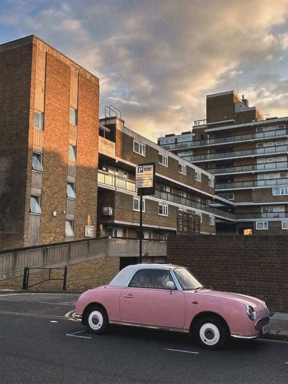 A small pink retro car is parked in front of a large, Brutalist-style brick apartment building under a cloudy sky. A parking…