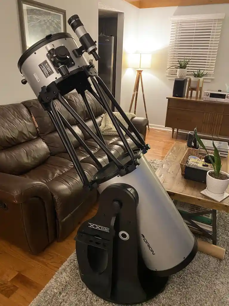 A large, silver and black Orion SkyQuest telescope is positioned indoors, leaning against a brown leather couch in a living…