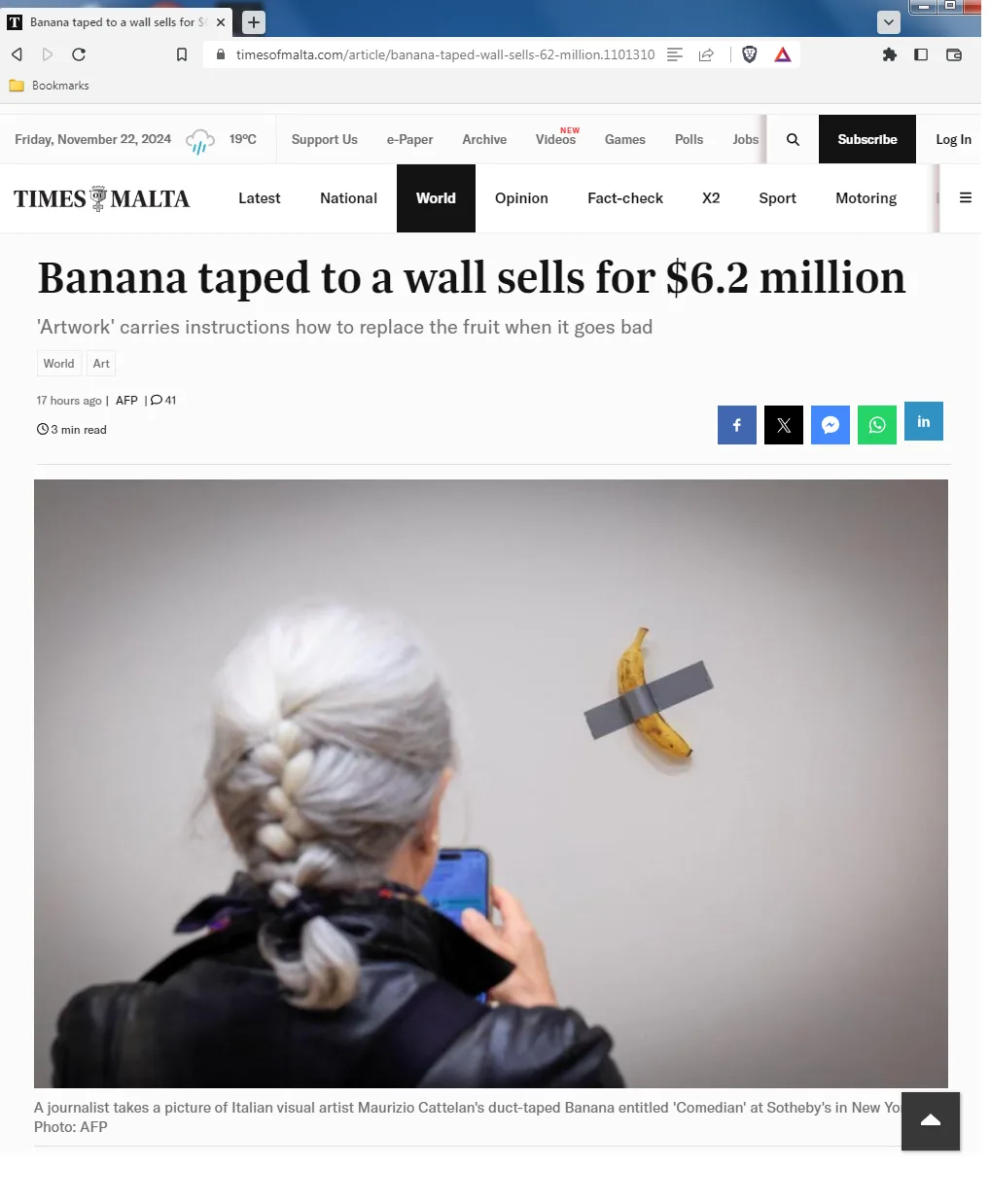 A person with white hair in a braid takes a photo of a single banana taped to a plain wall with duct tape. The image is a…