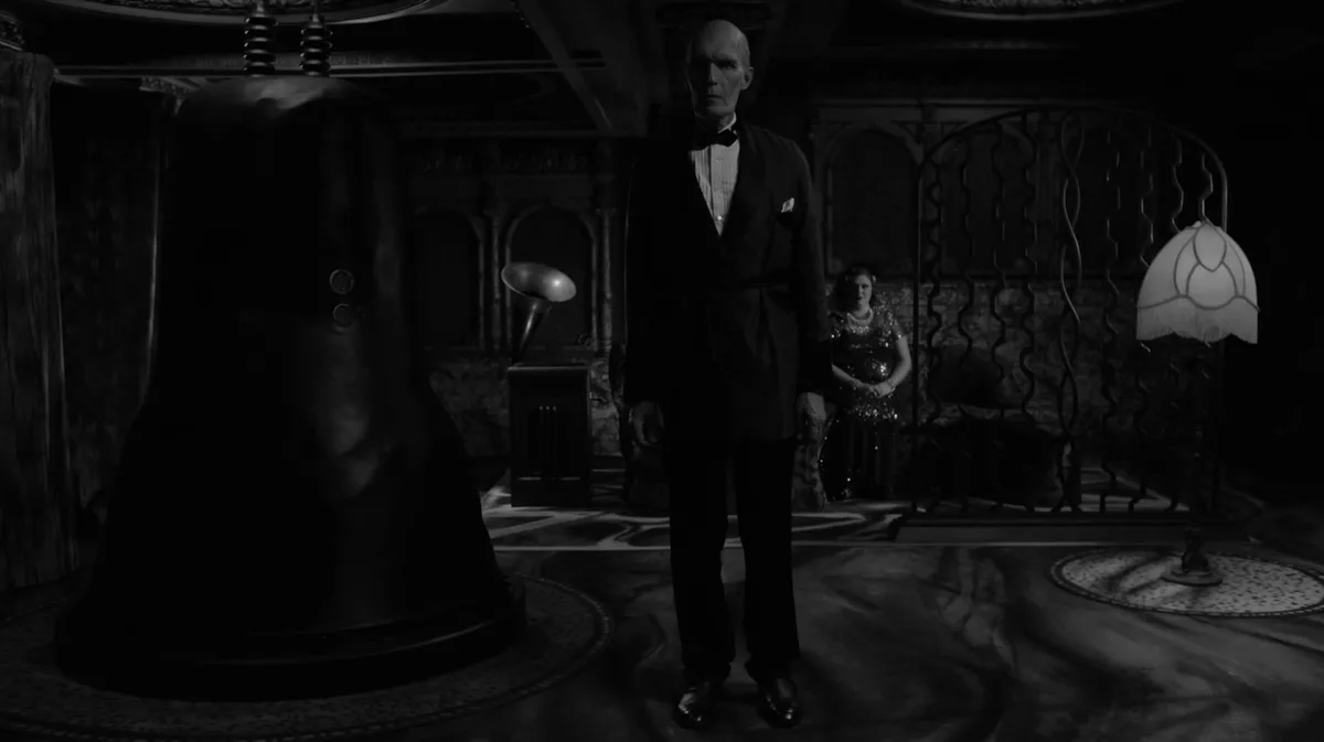 A black-and-white scene from the 1940 film 'The Invisible Man' shows a bald man in a tuxedo standing in a dimly lit, ornate…