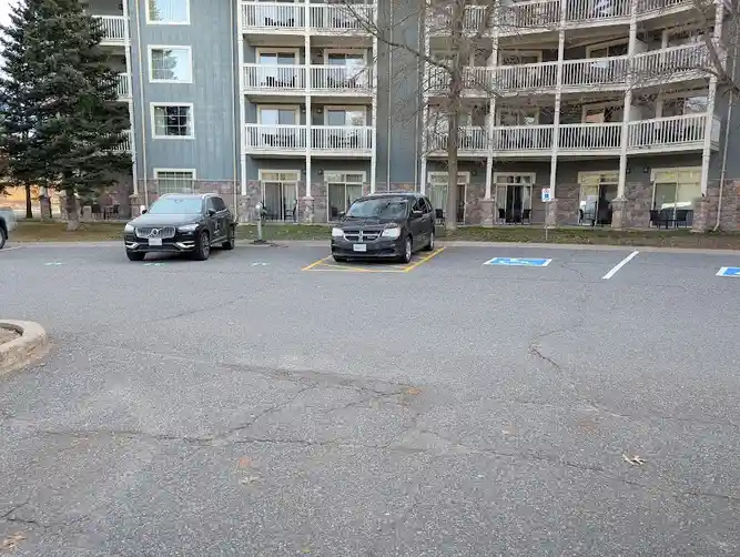 A parking lot with two cars parked in front of a multi-story apartment building. One car is parked in a clearly marked…