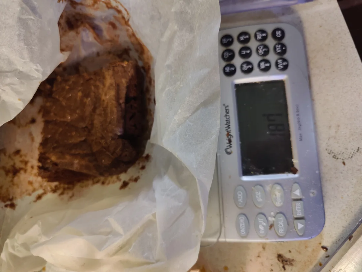A close-up photo shows a large, messy brownie resting on crumpled parchment paper next to a Weight Watchers brand food…