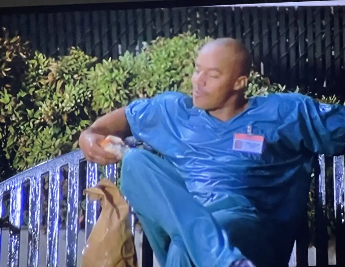 A man in a shiny blue rain suit sits on a wet metal bench at night, holding a snack, with green bushes and a black fence…