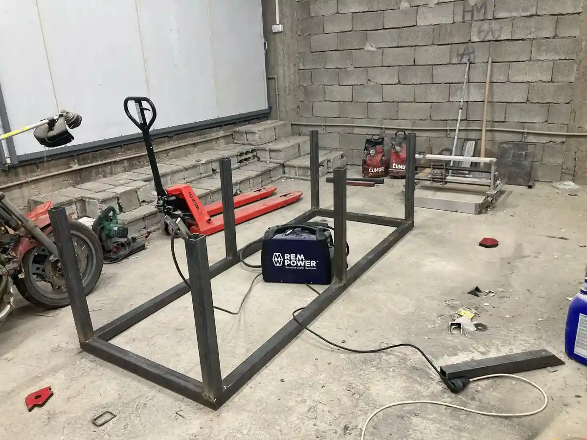 A workshop scene shows a partially assembled metal frame for a motorcycle, with welding equipment nearby. The frame is…