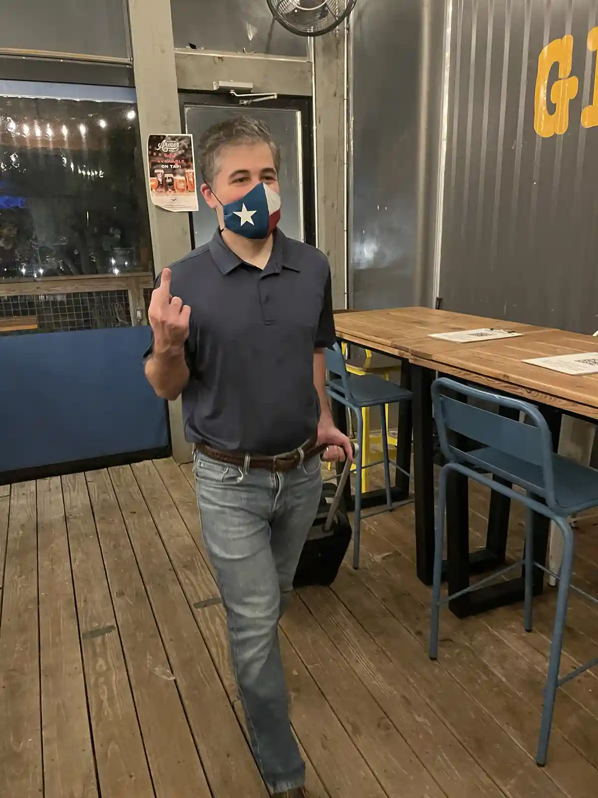 a man wearing a texas flag face mask and a dark polo shirt is walking indoors while holding a suitcase handle and making a…