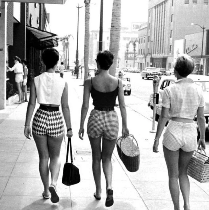 a black-and-white photo of three women walking away from the camera down a city sidewalk, wearing high-waisted shorts and…
