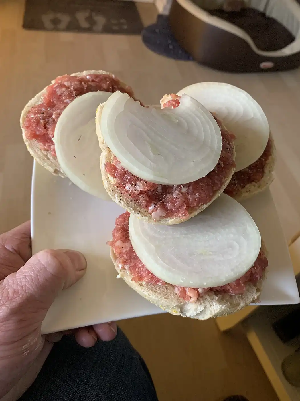 A hand holds a white plate with three open-faced burger buns topped with raw ground meat and large, thick slices of raw…