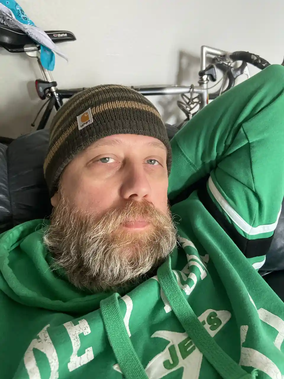 A man with a graying beard and a green hoodie lies on a couch, looking directly at the camera with a relaxed expression…